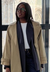 Beige trenchcoat i lager över en marinblå blazer med en vit topp, kombinerad med mörka jeans. Tjocka svarta glasögon ger en modern detalj.