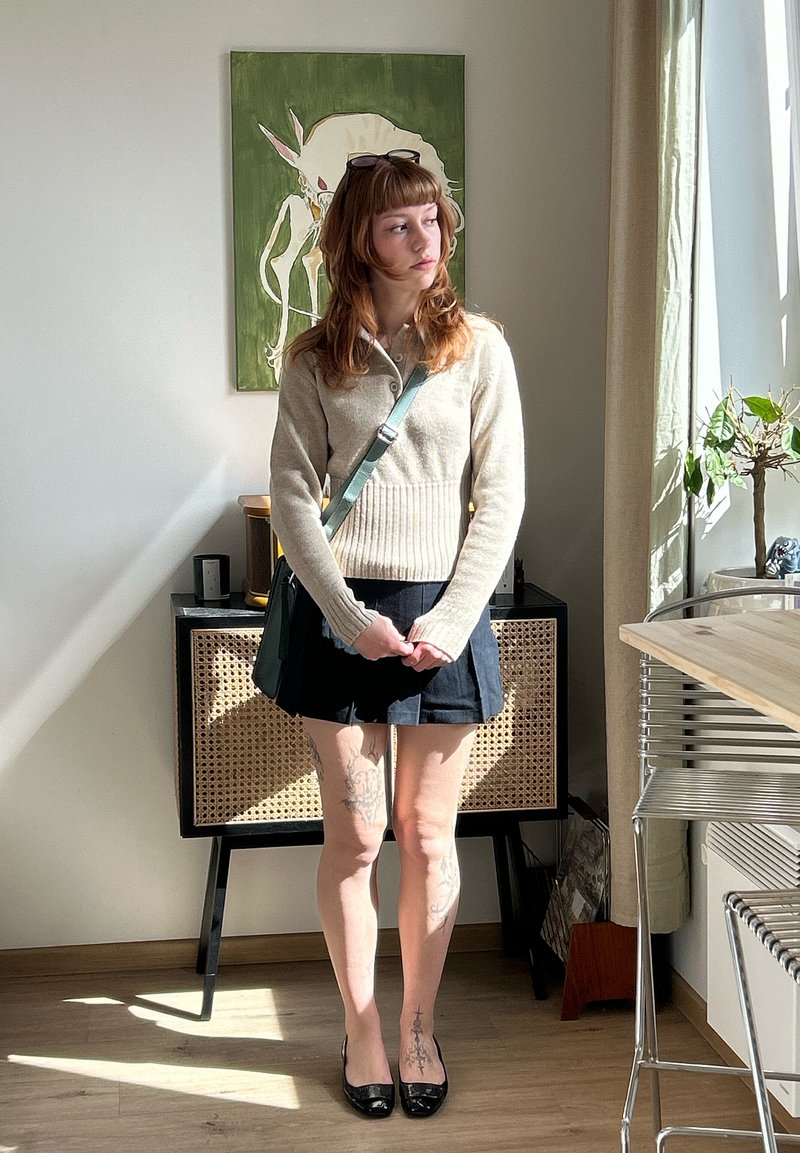 Jeune femme aux cheveux bruns, pull beige, jupe noire et tatouages sur les jambes se tient dans une pièce lumineuse près d'une armoire et d'une fenêtre avec une plante.