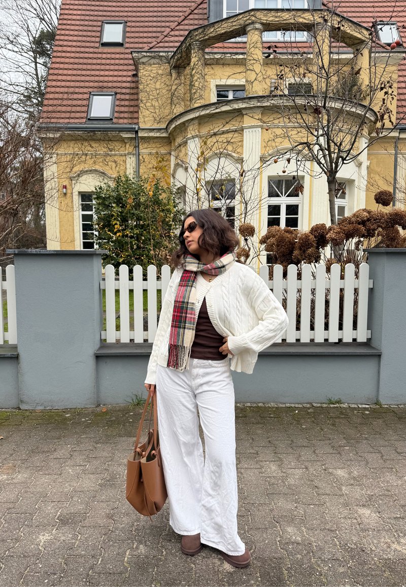 Cardigan alb cu model torsadat purtat peste un top maro, asortat cu pantaloni largi albi și ghete maro până la gleznă. Poartă o geantă tote maro.