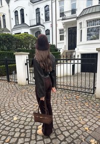 Femme aux longs cheveux bruns portant une veste sombre, un pantalon marron et un chapeau en fausse fourrure, se tient sur un chemin pavé en tenant un sac à main marron.