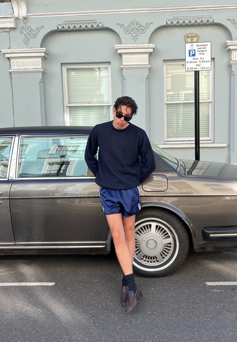 Homem a usar óculos de sol, pulôver azul marinho e calções azuis, encostado a um carro vintage castanho estacionado numa rua em frente a um edifício azul claro.