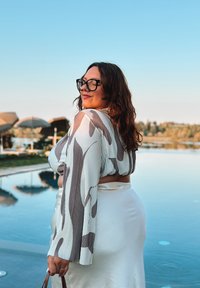 Blusa bianca con motivo astratto marrone e maniche lunghe, abbinata a una gonna aderente bianca, sullo sfondo di una piscina e cielo sereno.