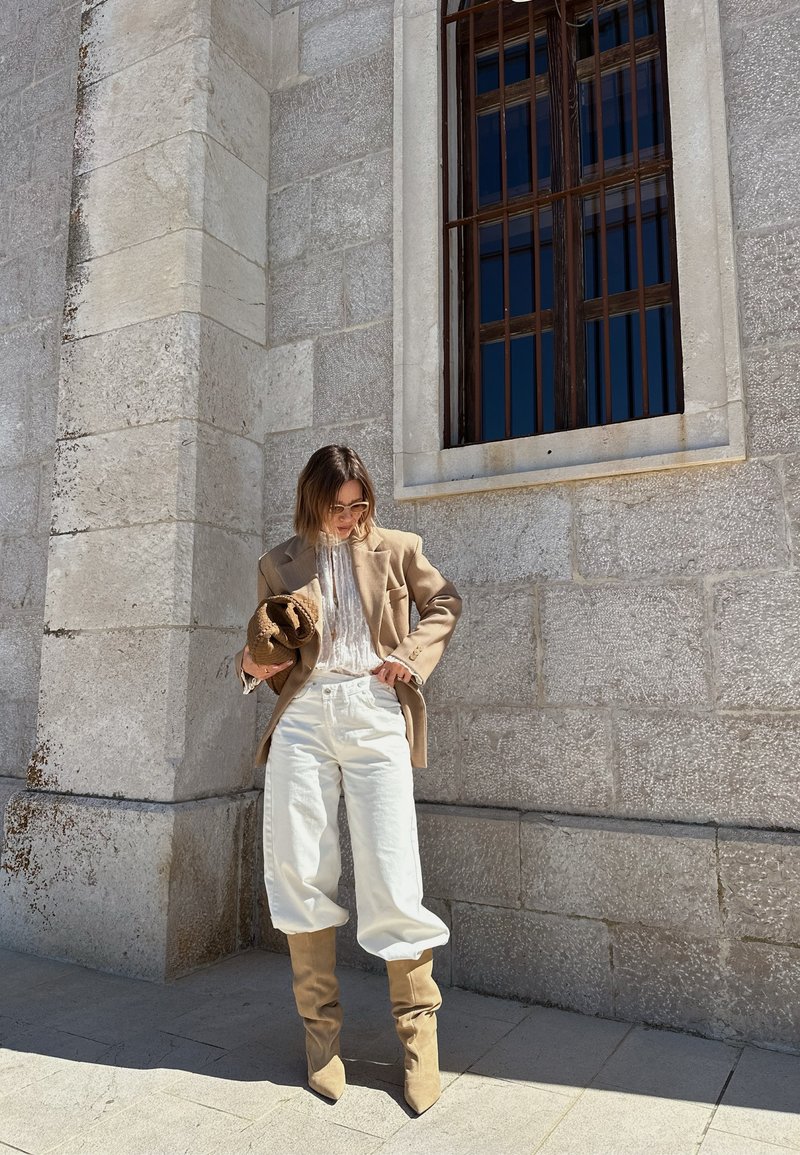 Femme en blazer beige, blouse blanche, pantalon blanc et bottes beige tenant un sac tissé, debout devant un bâtiment en pierre avec une fenêtre grillagée.