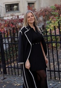 Black coat with white trim, over a ribbed black dress, paired with sheer black tights. The background features plants and a fence.