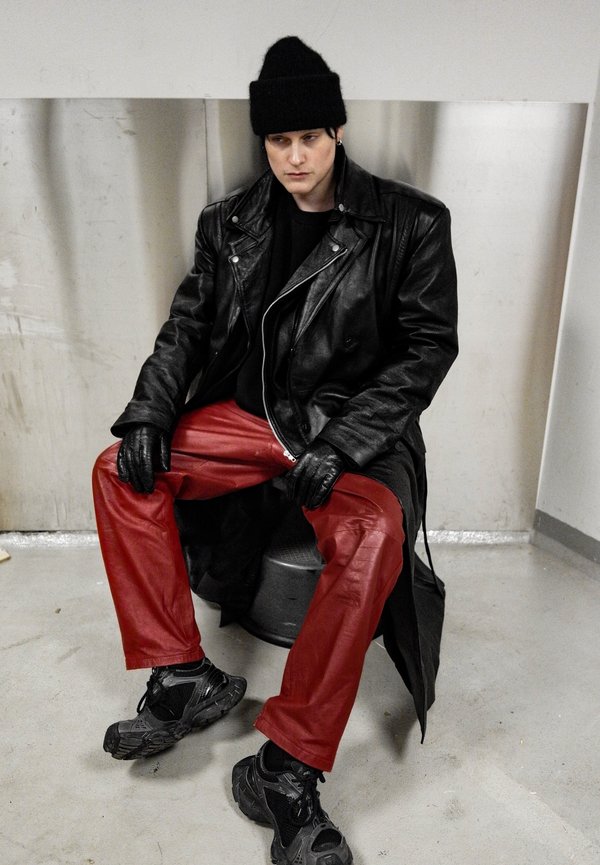 Chaqueta de cuero negra sobre una camiseta negra, combinada con pantalones de cuero rojo brillante y zapatillas negras. El look se completa con un gorro de punto y guantes desgastados.