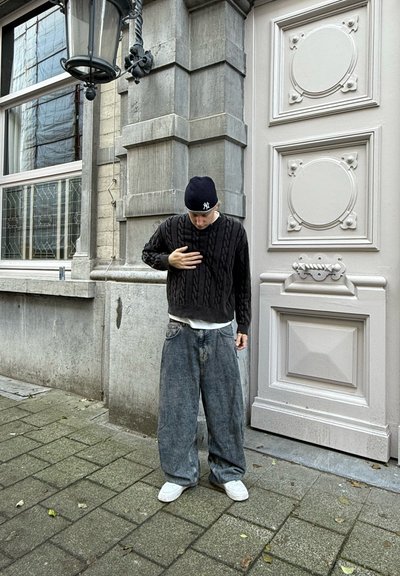 Suéter negro de punto con mangas largas, jeans de mezclilla azul sueltos y zapatillas blancas. De pie en una calle pavimentada cerca de un edificio.