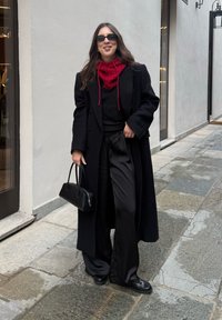 Čierny vlnený kabát cez čierny sveter, doplnený saténovými čiernymi nohavicami s širokými nohavicami. Červený šál s vzorom a malá čierna kabelka. Čierne slnečné okuliare.