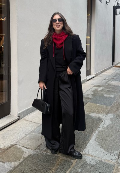 Abrigo de lana negra sobre un suéter negro, combinado con pantalones anchos de satén negros. Bufanda roja con estampado y un pequeño bolso negro. Gafas de sol negras.