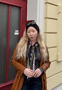 Hnědý kožený kabát, tmavý top, vzorovaný šátek v černé, bílé a zelené barvě. Dlouhé, vlnité blond vlasy s černou čelenkou. Pozadí s červenými dveřmi.