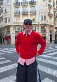 Maglione rosso lavorato a maglia, camicia a righe rosa sotto, pantaloncini larghi neri con strisce bianche, cappello grigio, occhiali da sole, in piedi su una striscia pedonale.