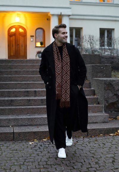 Abrigo negro, bufanda marrón con un diseño geométrico blanco, ropa negra y zapatillas blancas. Escalones que llevan a un edificio en el fondo.