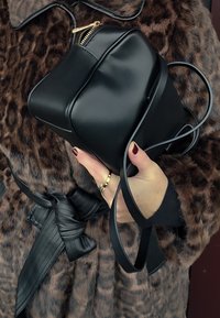 Sac à main en cuir noir de forme rectangulaire, avec un détail de fermeture éclair dorée et une longue lanière fine, tenu à la main sur un fond à motif léopard.