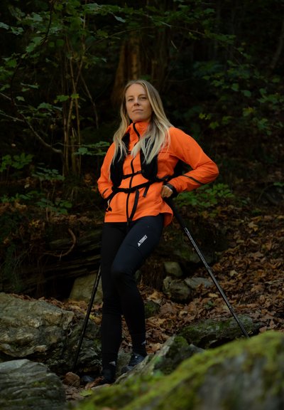 Chaqueta impermeable naranja brillante con un arnés negro, pantalones negros ajustados y bastones de senderismo, con un fondo boscoso y rocoso.