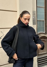 Námornícka modrá fleecová bunda s vysokým golierom a širokými rukávmi. Spárovaná s čiernou koženou tašku a tmavými džínsami, postavená proti béžovej stene.