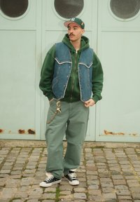 Veste en jean avec bords effilochés portée sur un sweat à capuche vert, associée à un pantalon vert ample et des baskets noires ; porte-clés accroché au pantalon.