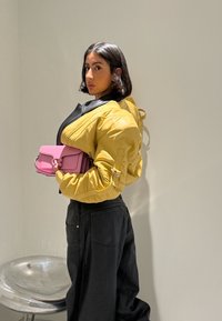 Femme aux cheveux mi-longs et foncés, portant une veste jaune et un pantalon noir, tenant un sac à main rose, debout à côté d'une chaise argentée.