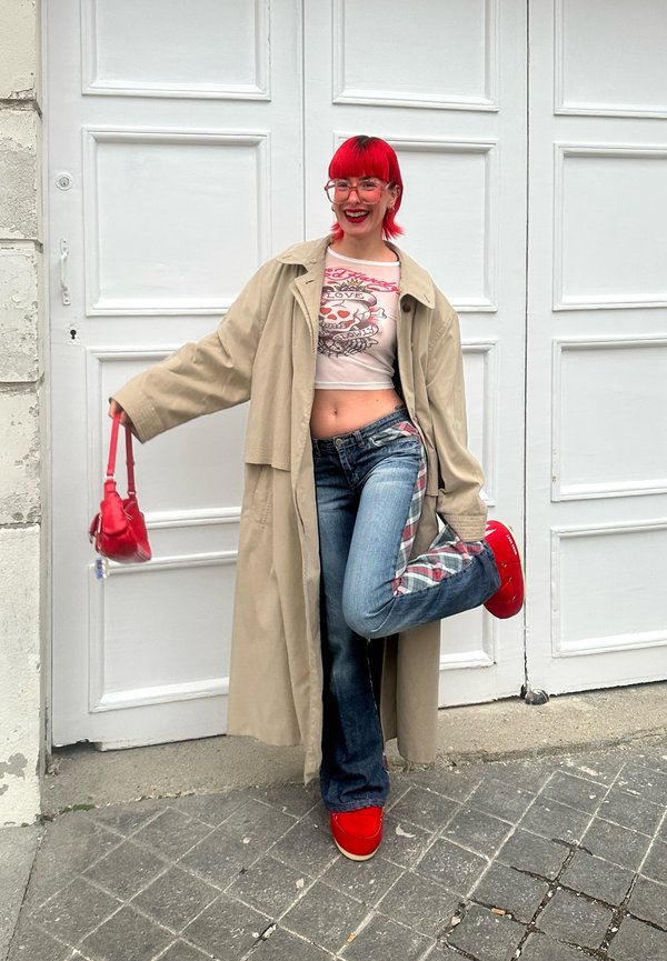 Cappotto trench beige sopra una maglietta crop bianca con grafica, jeans blu con dettagli in tartan e scarpe e borsa di colore rosso brillante.