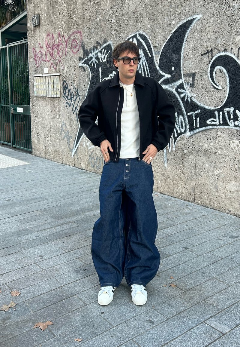 Pantalon en denim bleu surdimensionné avec une taille haute et des boutons à l'avant, associé à une chemise blanche et une veste bomber noire. Baskets neutres.