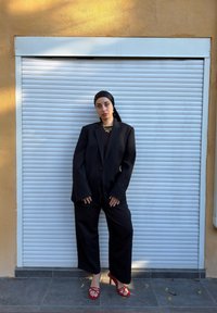 Črna prevelika suknjič in široke hlače z visokim pasom, v kombinaciji z rdečimi sandali z naramnicami. Videz dopolnjuje temna majica in večplastni zlati nakit.