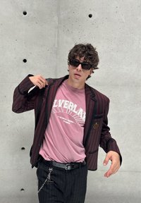 Rdeča in črna prugasta blazer jakna z insignijo, roza grafična majica, črne pruge hlače, sončna očala in detajl s verigo. Tekstura materiala.