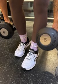 Topánky Reebok Nano XV v bielej farbe s čiernymi akcentmi, nosené s ružovými ponožkami, počas držania 10 kg činkov Eleiko na podlahe posilňovne.