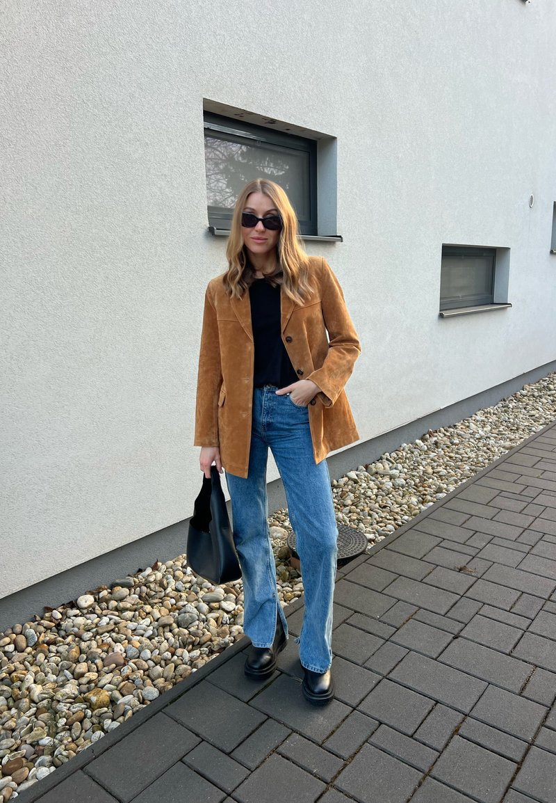 Frau trägt ein braunes Wildleder-Blazer, ein schwarzes Oberteil, blaue Jeans, schwarze Stiefel und eine Sonnenbrille, steht auf einem gepflasterten Weg neben einer mit Kieselsteinen gesäumten Mauer.