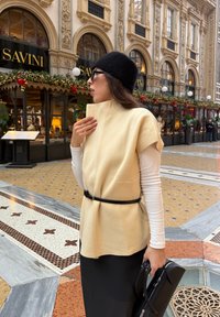 Beige sleeveless top with a high collar, belted with a black strap, over a white long-sleeve shirt. Square black handbag, patterned flooring.
