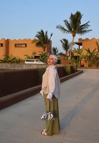 Ένα άτομο φοράει ένα λευκό πουκάμισο με μακριά μανίκια, μια μπεζ κασκόλ και πράσινο παντελόνι φαρδύ, κρατώντας μια ασημένια τσάντα με μαύρες βούλες, στέκεται σε έναν διάδρομο.