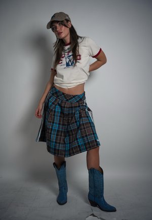 Mujer con camiseta gráfica blanca, falda de cuadros azules, botas vaqueras azules y gorra de cuadros, de pie frente a un fondo gris liso.