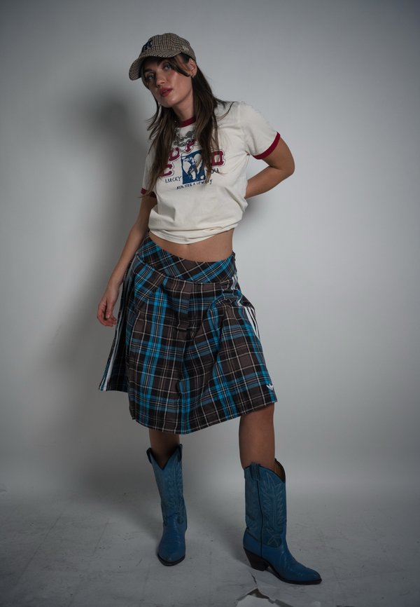 Vrouw draagt een wit T-shirt met grafische opdruk, een blauw geruite rok, blauwe cowboylaarzen en een geruit petje, staand tegen een effen grijze achtergrond.