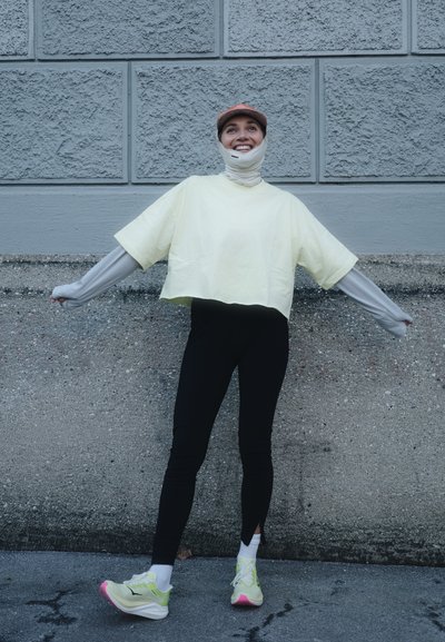 Persona con camisetas de manga larga y corta superpuestas, mallas negras, zapatillas de neón amarillo y una gorra, de pie con los brazos extendidos contra una pared gris.