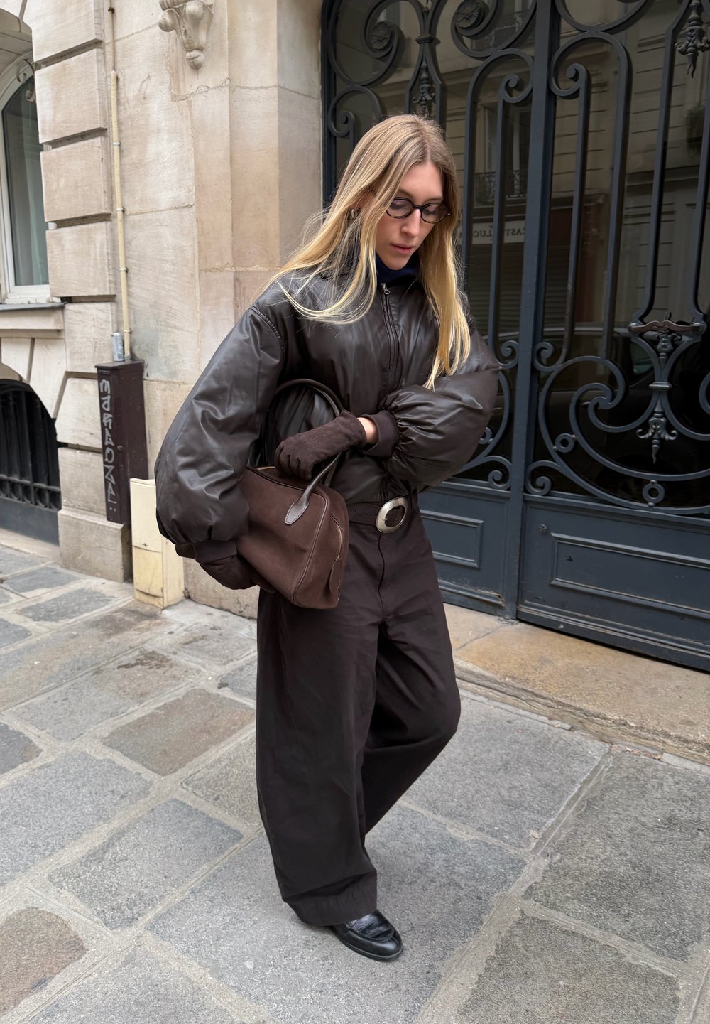 Braune, wattierte Bomberjacke, strukturierte braune Hose, schwarze Handschuhe und eine braune Tasche. Trägt eine Brille, geht auf einer steinernen Straße mit einer dekorativen Tür.