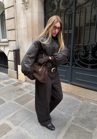 Braune, wattierte Bomberjacke, strukturierte braune Hose, schwarze Handschuhe und eine braune Tasche. Trägt eine Brille, geht auf einer steinernen Straße mit einer dekorativen Tür.