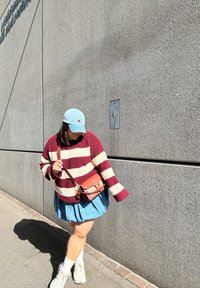Maglione a righe bordeaux e crema, gonna di jeans, cappello azzurro chiaro, borsa marrone, calzini bianchi e sneakers chiare davanti a un muro grigio testurizzato.
