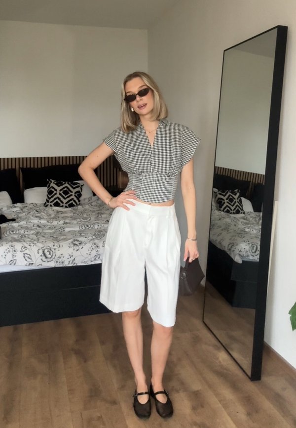 Femme portant des lunettes de soleil, un haut à carreaux noir et blanc, un short blanc jusqu'aux genoux et des chaussures noires se tient dans une chambre à côté d'un miroir en pied.