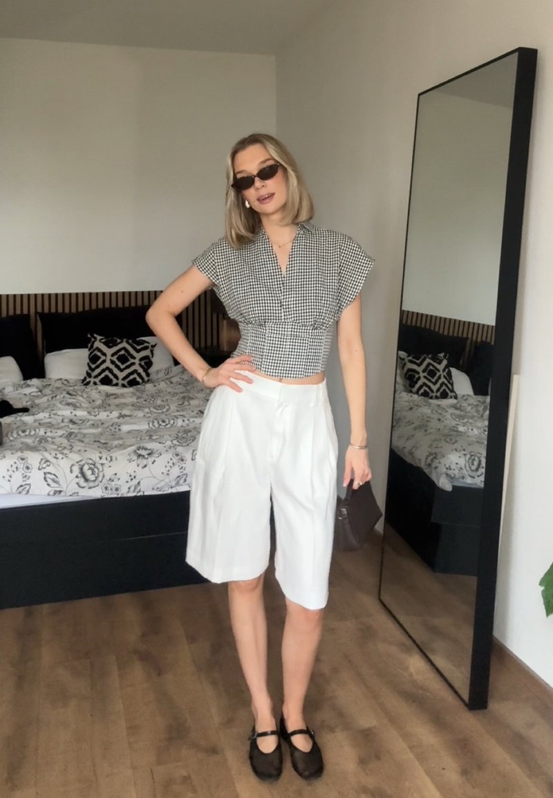 Femme portant des lunettes de soleil, un haut à carreaux noir et blanc, un short blanc jusqu'aux genoux et des chaussures noires se tient dans une chambre à côté d'un miroir en pied.