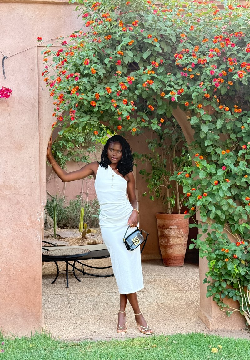 Robe blanche sans manches avec texture plissée, se tenant dans une arche entourée de feuillage vert vif et de fleurs orange. Sac à main noir avec des accents dorés.