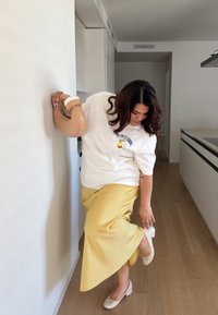 Egy fehér pólót és sárga selyem szoknyát viselő személy áll egy fal mellett, kerek formájú, világosbarna táskát tartva a kezében.