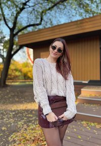 Kabelstrick-Cremepullover, bordeauxfarbenen Lack-Minirock und eine kleine passende Handtasche. Außenbereich mit herbstlichem Laub und Holzstufen.