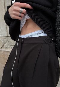 Zwarte broek met plooien, een wit shirt eronder en een zichtbare Calvin Klein tailleband. Zilveren ringen worden om de vingers gedragen.