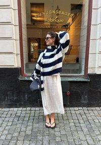 Fleckiger gestreifter Pullover in Marineblau und Weiß, knielanger weißer Leinenrock, schwarze Flip-Flops, hält eine graue Tasche, steht auf Kopfsteinpflaster.