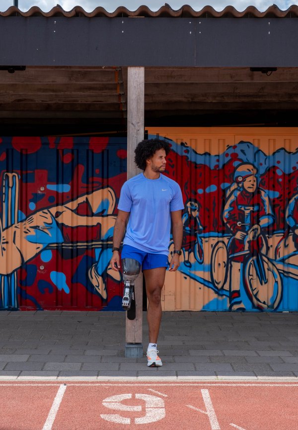 Uomo con una gamba protesica in abbigliamento sportivo appoggiato a un palo su una pista, murale sportiva colorata sul muro dietro di lui.