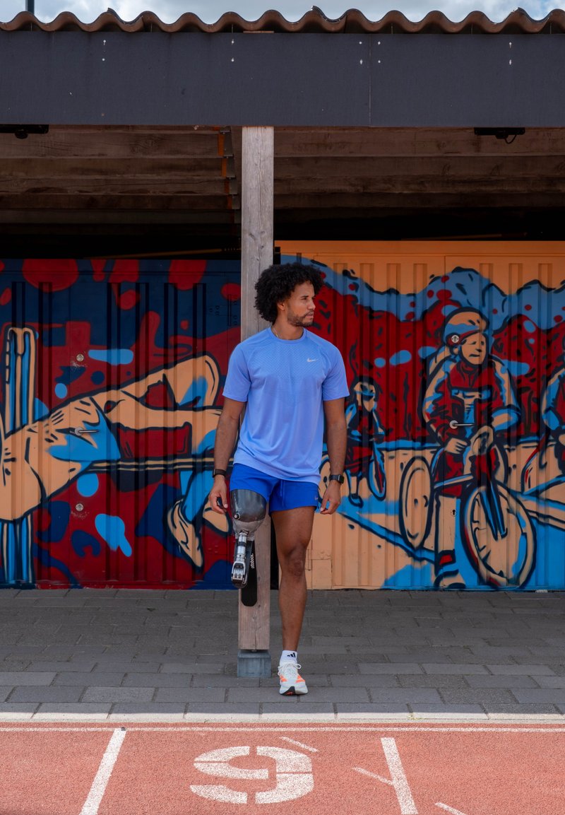 Uomo con una gamba protesica in abbigliamento sportivo appoggiato a un palo su una pista, murale sportiva colorata sul muro dietro di lui.