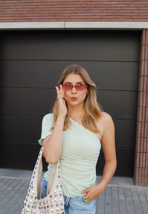 Top verde menta de un solo hombro, de tela ligera; gafas de sol redondas rosas vintage; bolso beige con tachuelas plateadas; pantalones cortos de mezclilla casuales; cabello ondulado y claro.