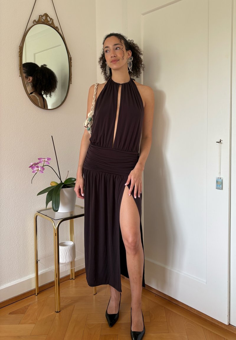 Femme en robe sombre sans manches avec une fente avant et des boucles d'oreilles imposantes se tient à côté d'une petite table avec une orchidée en pot et un miroir mural.