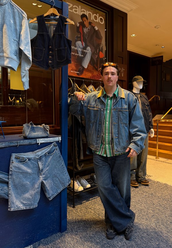 Joven con chaqueta vaquera y jeans se apoya en un perchero en una tienda con ropa vaquera y un maniquí al fondo.