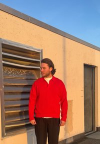 Κόκκινο ριγωτό πουλόβερ με γιακά, φορεμένο πάνω από ένα λευκό πουκάμισο. Το άτομο στέκεται δίπλα σε ένα μεταλλικό αεραγωγό μπροστά σε έναν μπεζ τοίχο.