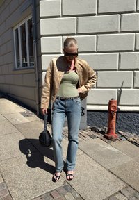 Casaco bomber de couro bege com detalhe em texto, top de alças verde e riscas, jeans azul claro, bolsa preta e sandálias pretas. Fundo urbano.