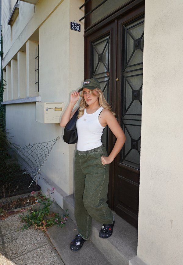 Joven mujer con camiseta sin mangas blanca, pantalones verdes y gorra está de pie fuera de la puerta de un edificio con el número 25a, ajustándose la gorra con una mano.