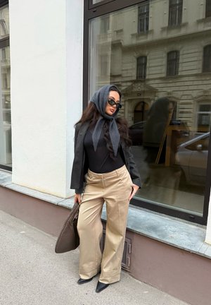 Mujer con blusa negra, pañuelo gris en la cabeza, chaqueta gris oscuro, pantalones anchos beige y zapatos negros puntiagudos, apoyada contra la ventana de un edificio.